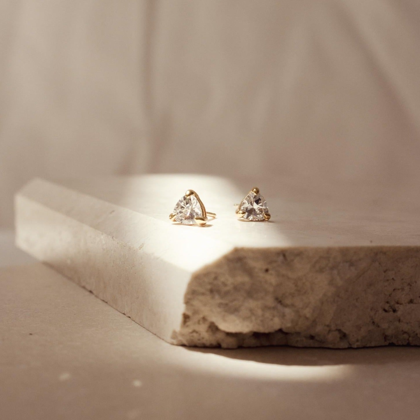 Triangle Diamond Studs rest on a textured stone slab in soft, natural light, showcasing their classic elegance and timeless style.
