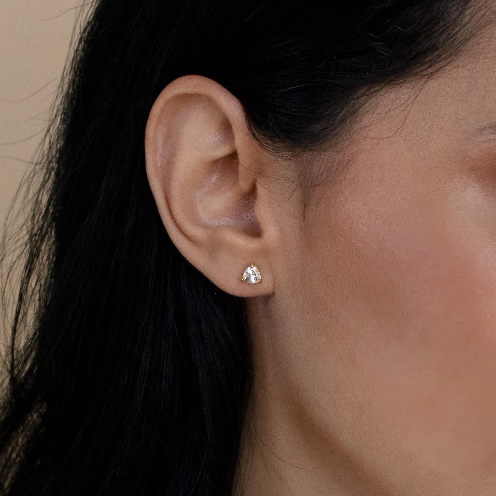 Close-up of an ear adorned with Triangle Diamond Studs—small, gold earrings in a geometric shape—offering an elegant, modern twist on classic jewelry against a neutral background.