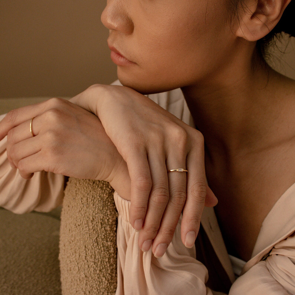 A woman rests her chin on her hands, highlighting a slim Baguette Diamond Ring and gold rings, styled with a beige blouse against a neutral backdrop.