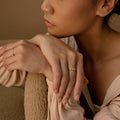 A woman rests her chin on her hands, highlighting a slim Baguette Diamond Ring and gold rings, styled with a beige blouse against a neutral backdrop.