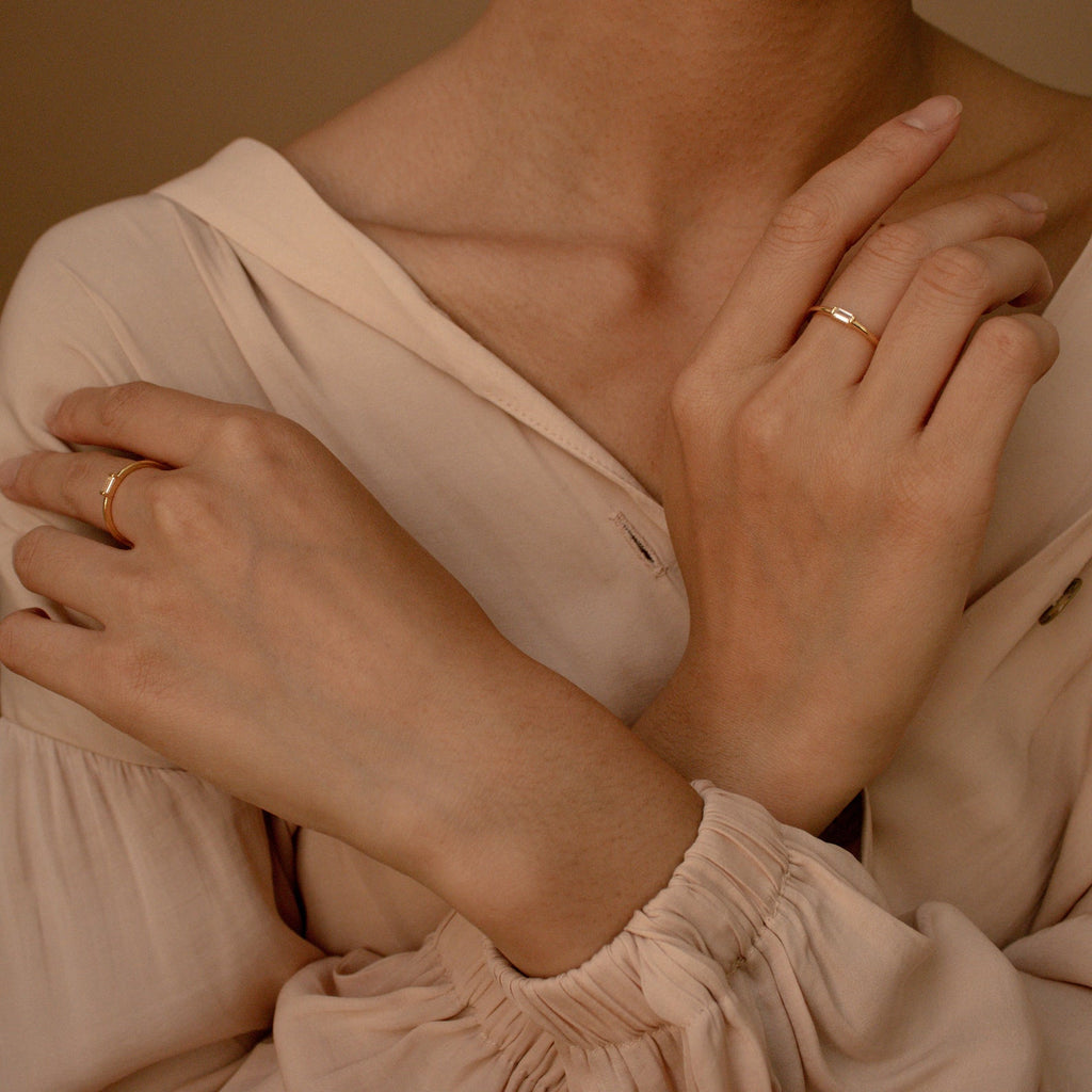 A person in a cream blouse crosses their arms, showing two gold rings on their fingers: a slim band and the Baguette Diamond Ring in an elegant east-west setting.