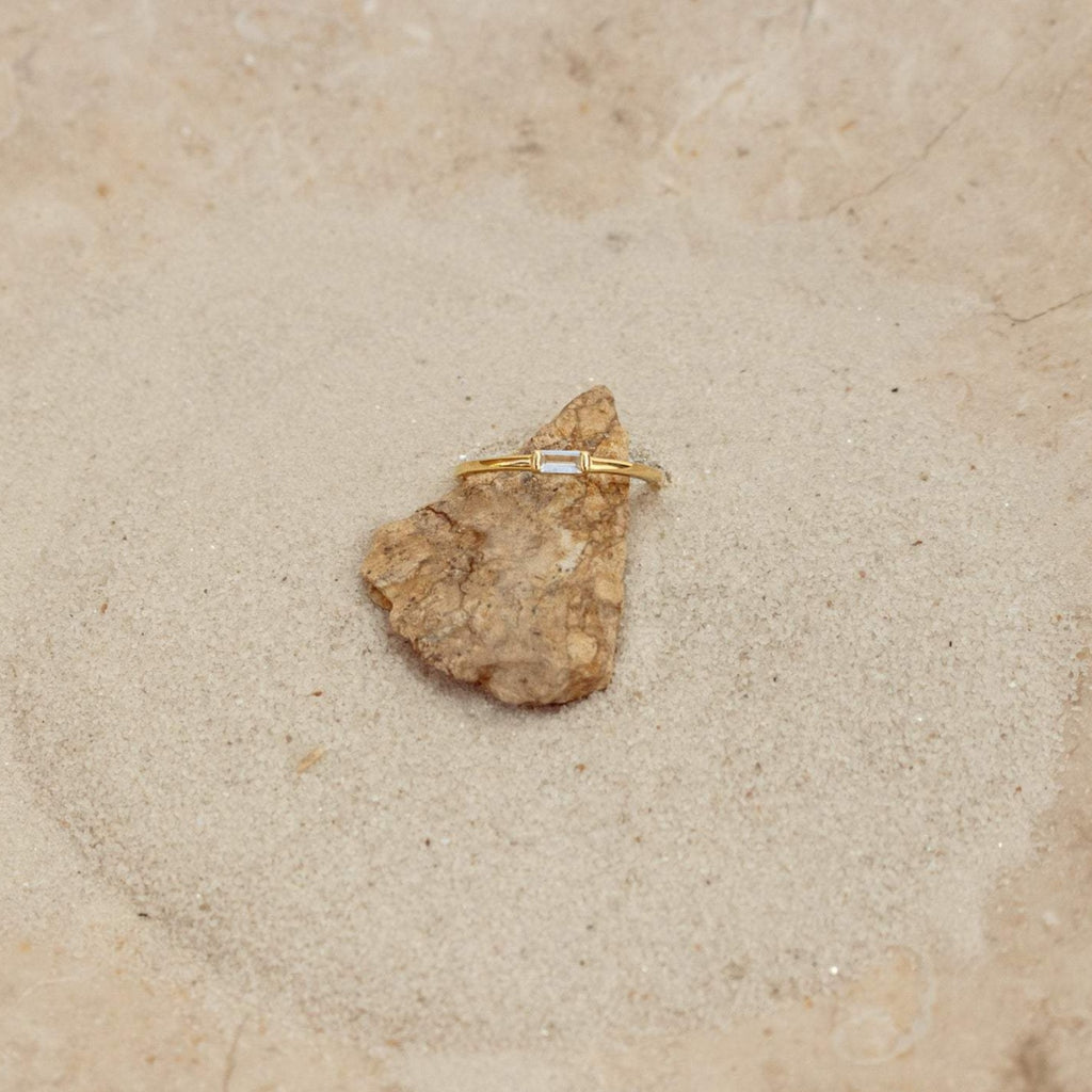 The Baguette Diamond Ring features a slim gold band and baguette diamonds, displayed on a small rock set on sand with a stone background.