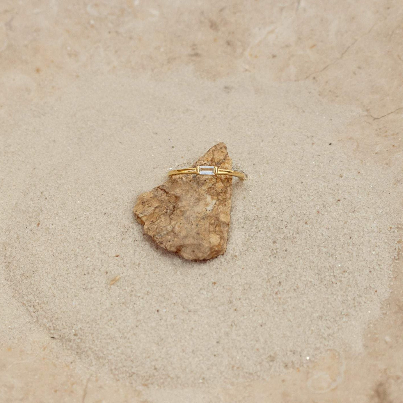 The Baguette Diamond Ring features a slim gold band and baguette diamonds, displayed on a small rock set on sand with a stone background.