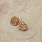 Two Baguette Diamond Rings with rectangular stones in an east-west setting rest delicately on small rocks atop a sandy, textured surface.