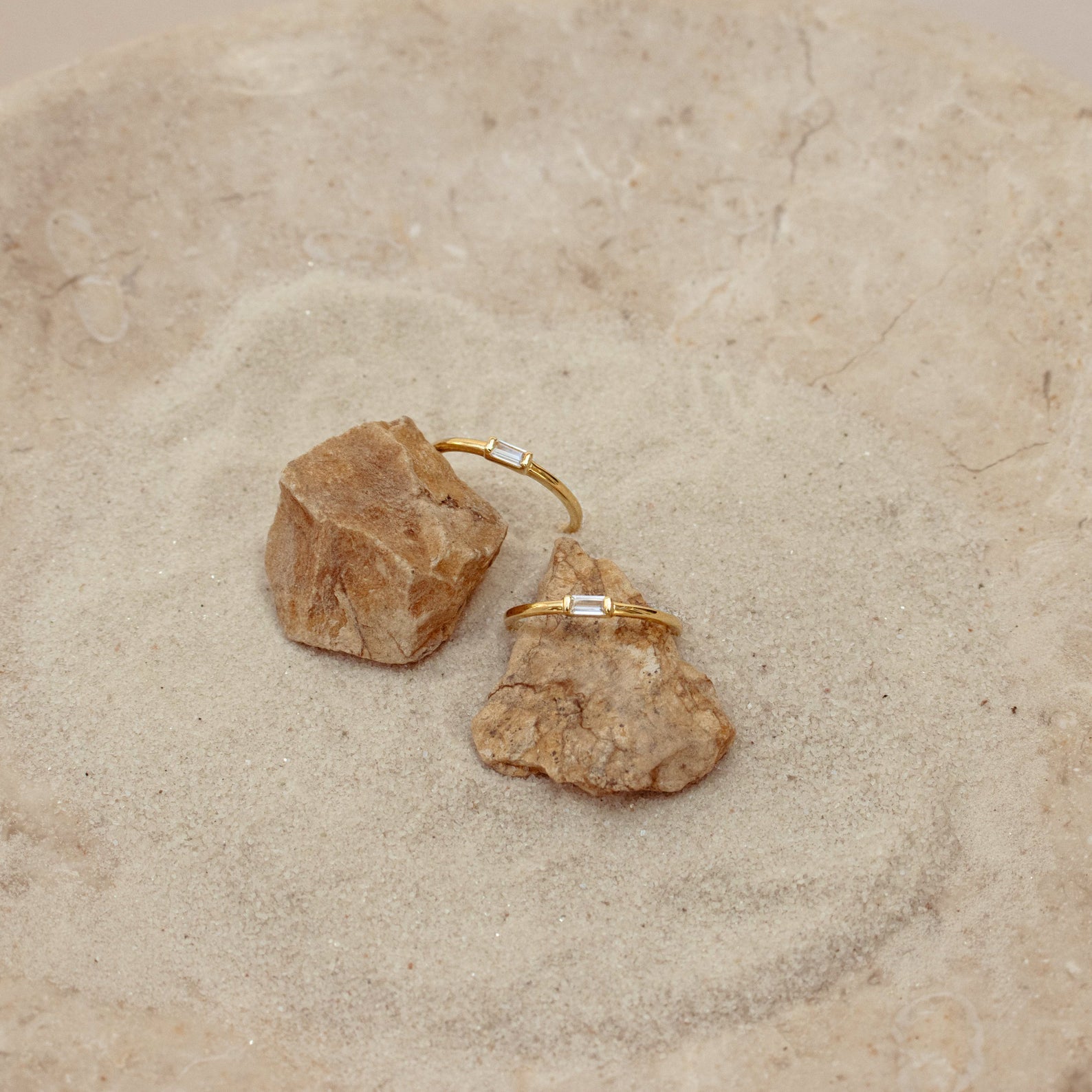 Two Baguette Diamond Rings with rectangular stones in an east-west setting rest delicately on small rocks atop a sandy, textured surface.