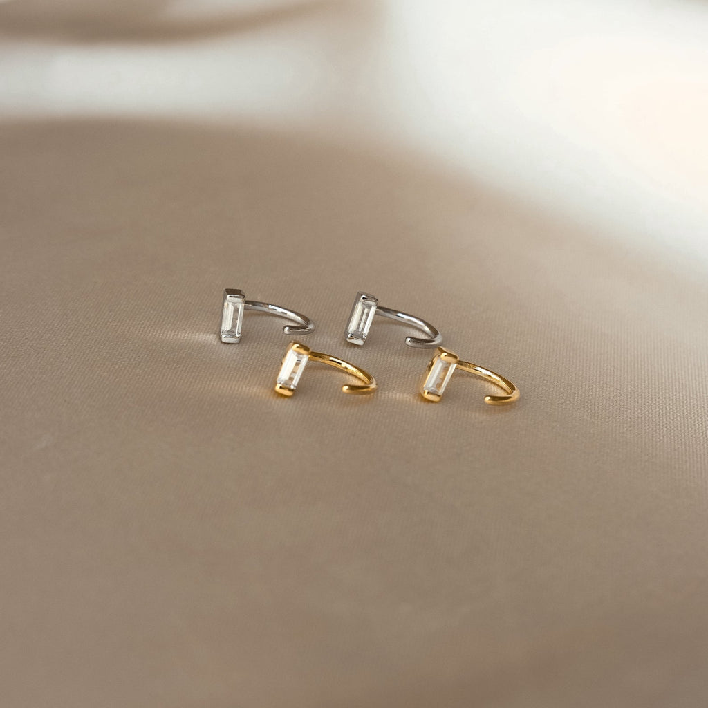 Shayla Baguette Huggies, featuring two gold and two silver open-ended rings with baguette-shaped stones, are displayed on a beige surface.
