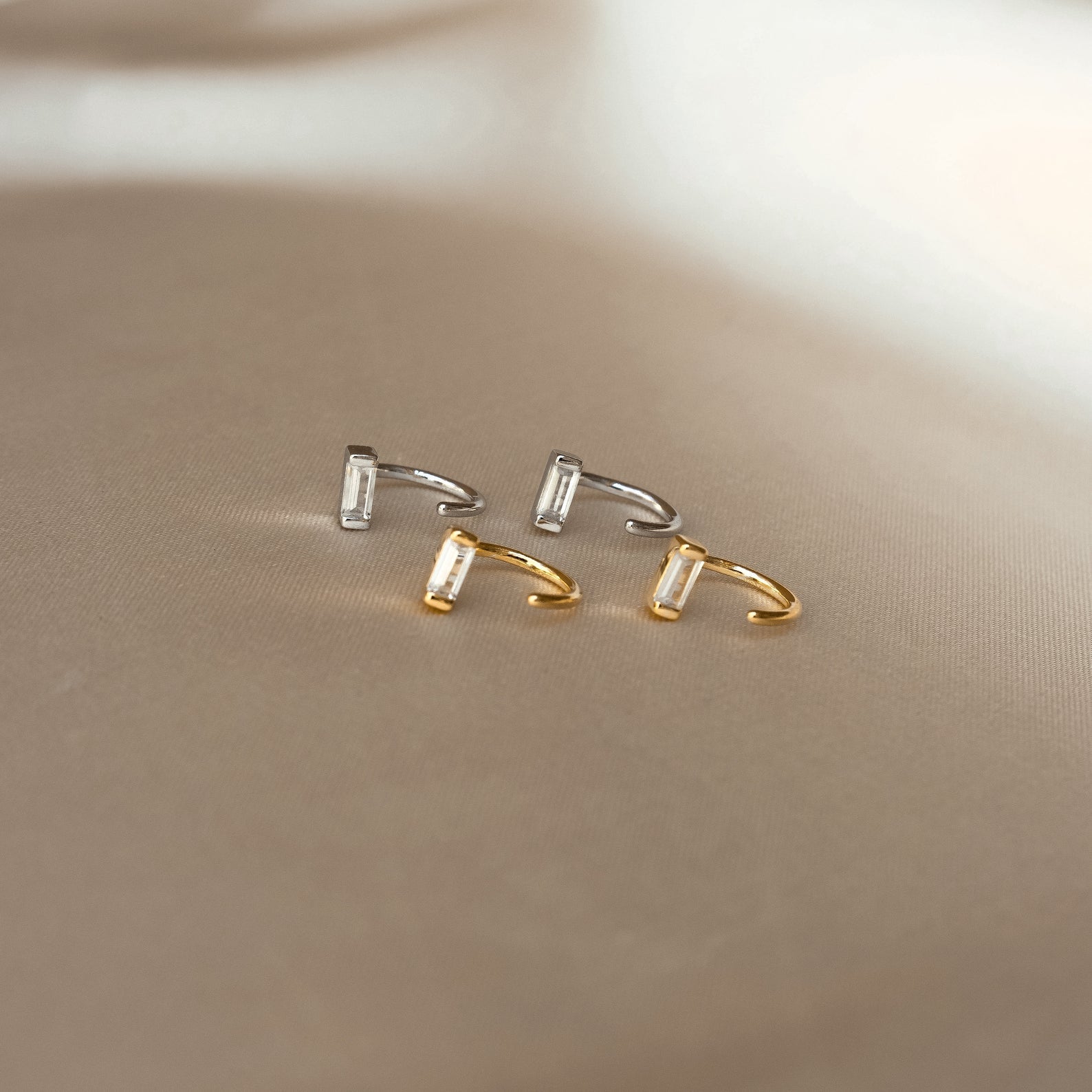 Shayla Baguette Huggies, featuring two gold and two silver open-ended rings with baguette-shaped stones, are displayed on a beige surface.