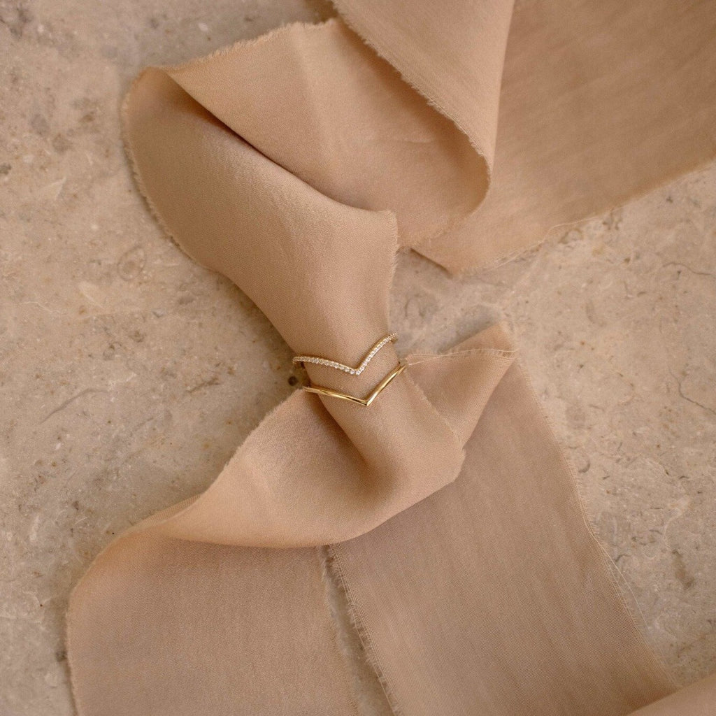 The Curve Stacking Set, featuring two gold and diamond chevron rings, is elegantly displayed on beige fabric atop a stone surface.