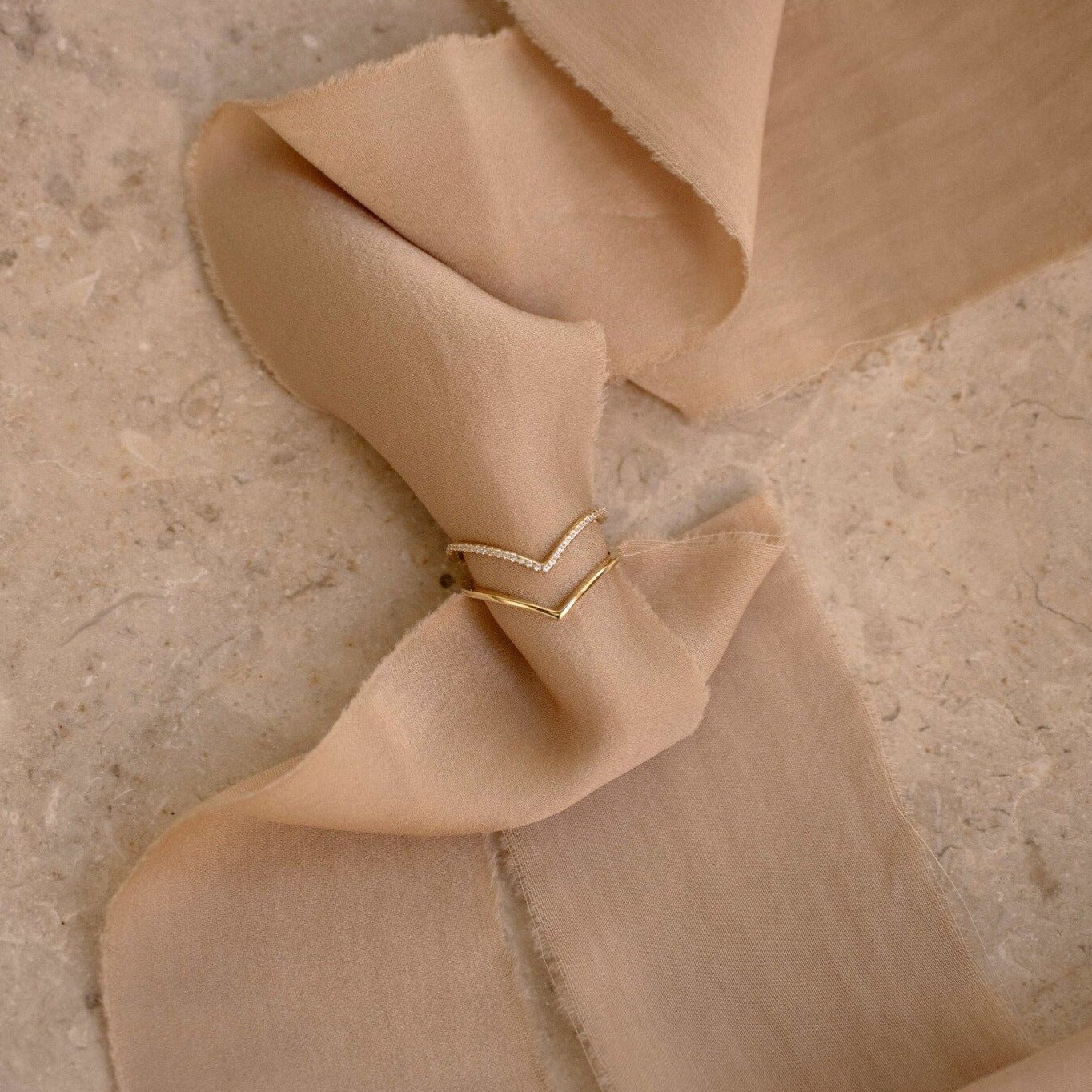 The Curve Stacking Set, featuring two gold and diamond chevron rings, is elegantly displayed on beige fabric atop a stone surface.