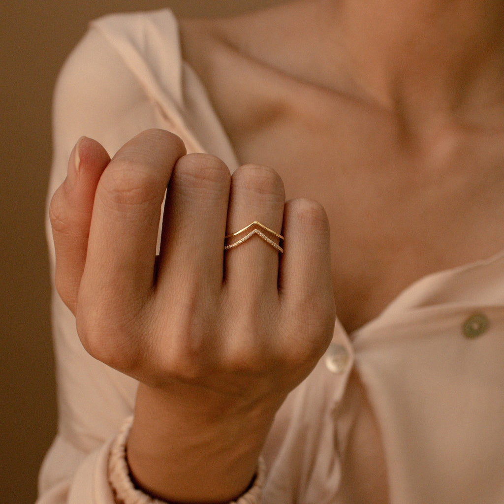 A hand showcases the Curve Stacking Set—two gold chevron rings styled together for a chic look, paired with a cream off-shoulder blouse.