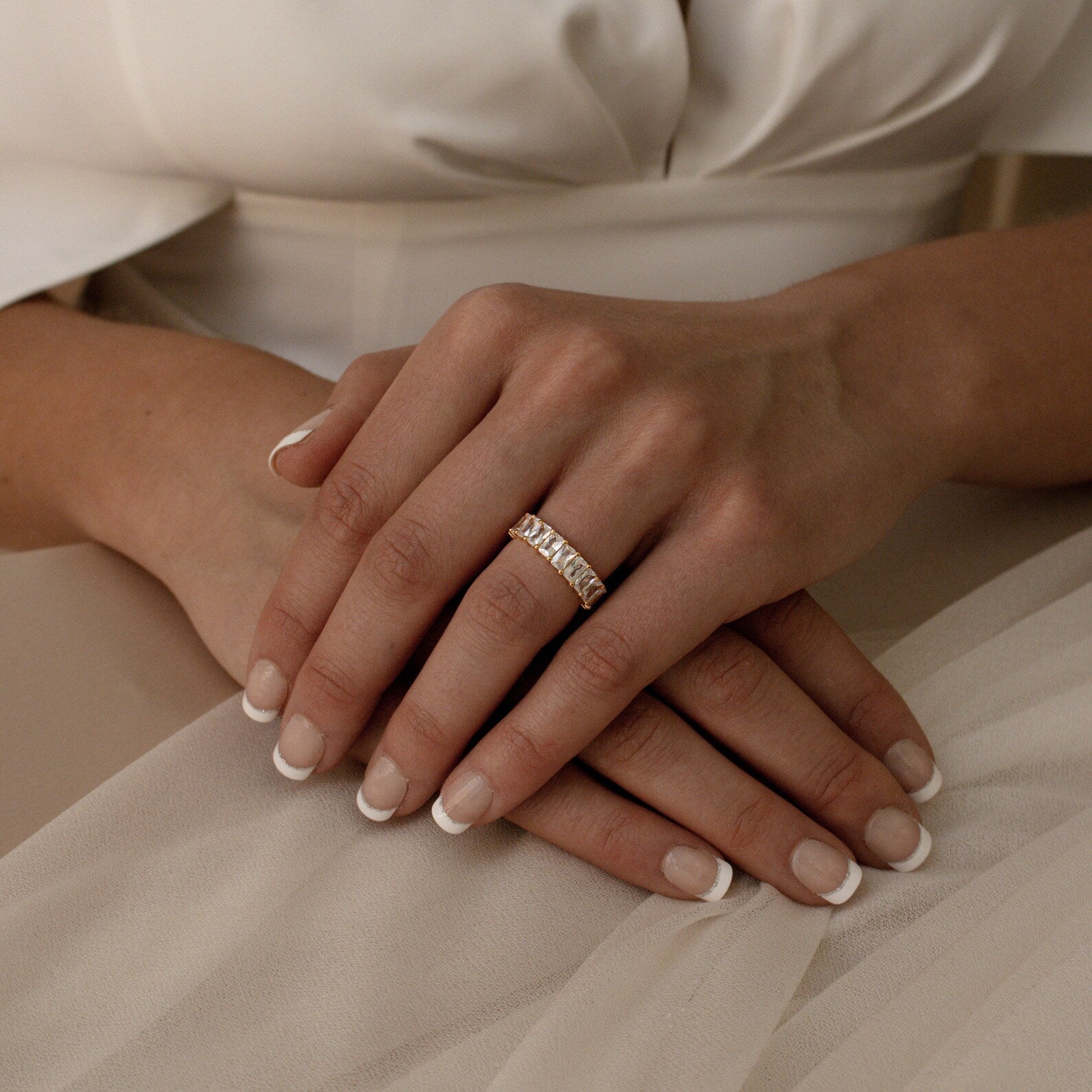 A woman's hands with a French manicure, wearing the Colette Eternity Ring—a gold statement ring with baguette diamonds—resting on a white fabric dress.