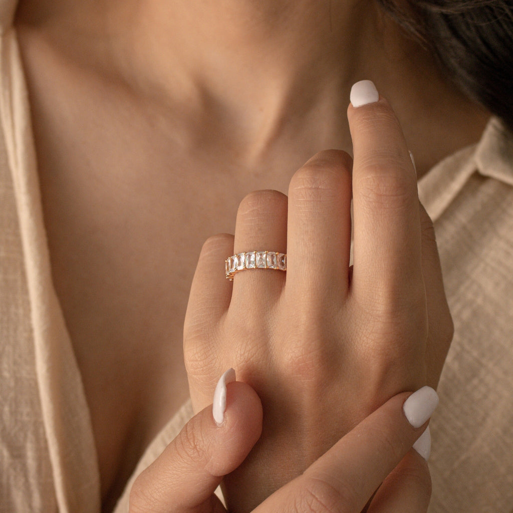 A woman with light nail polish wears the Colette Eternity Ring—a gold statement piece adorned with rectangular baguette diamonds—on her ring finger.