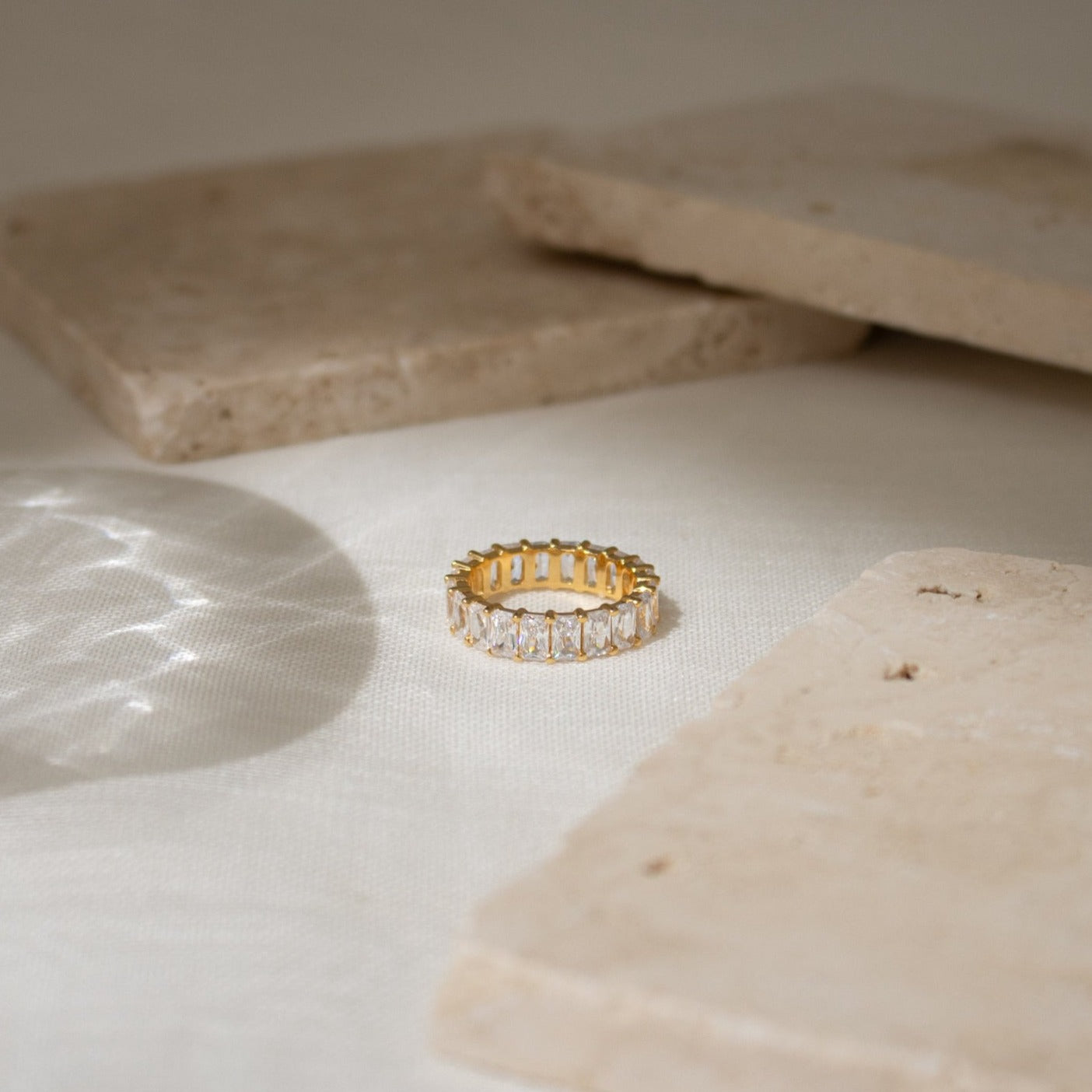 The Colette Eternity Ring, crafted in gold with rectangular baguette diamonds, rests on a light textured surface surrounded by stone slabs.