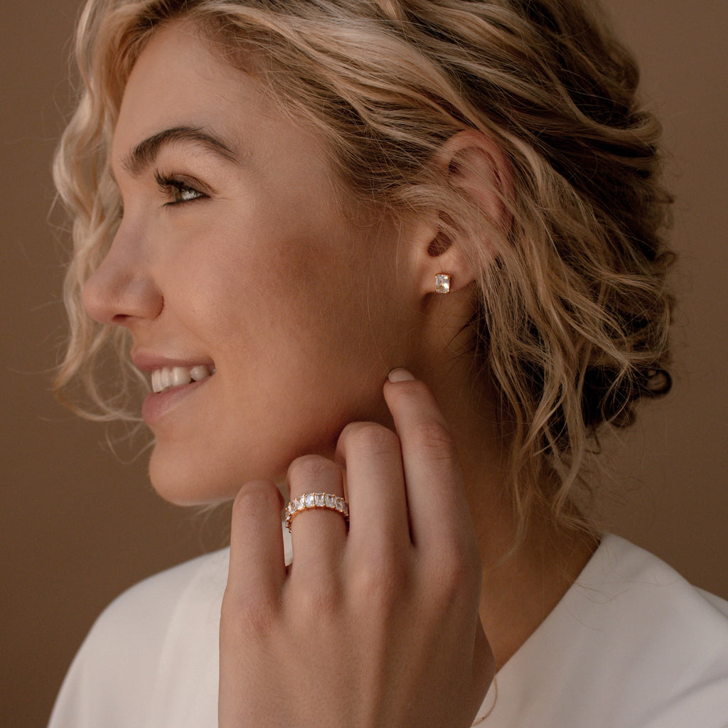 A smiling woman with curly blonde hair, wearing earrings and the Colette Eternity Ring, gently touches her face.