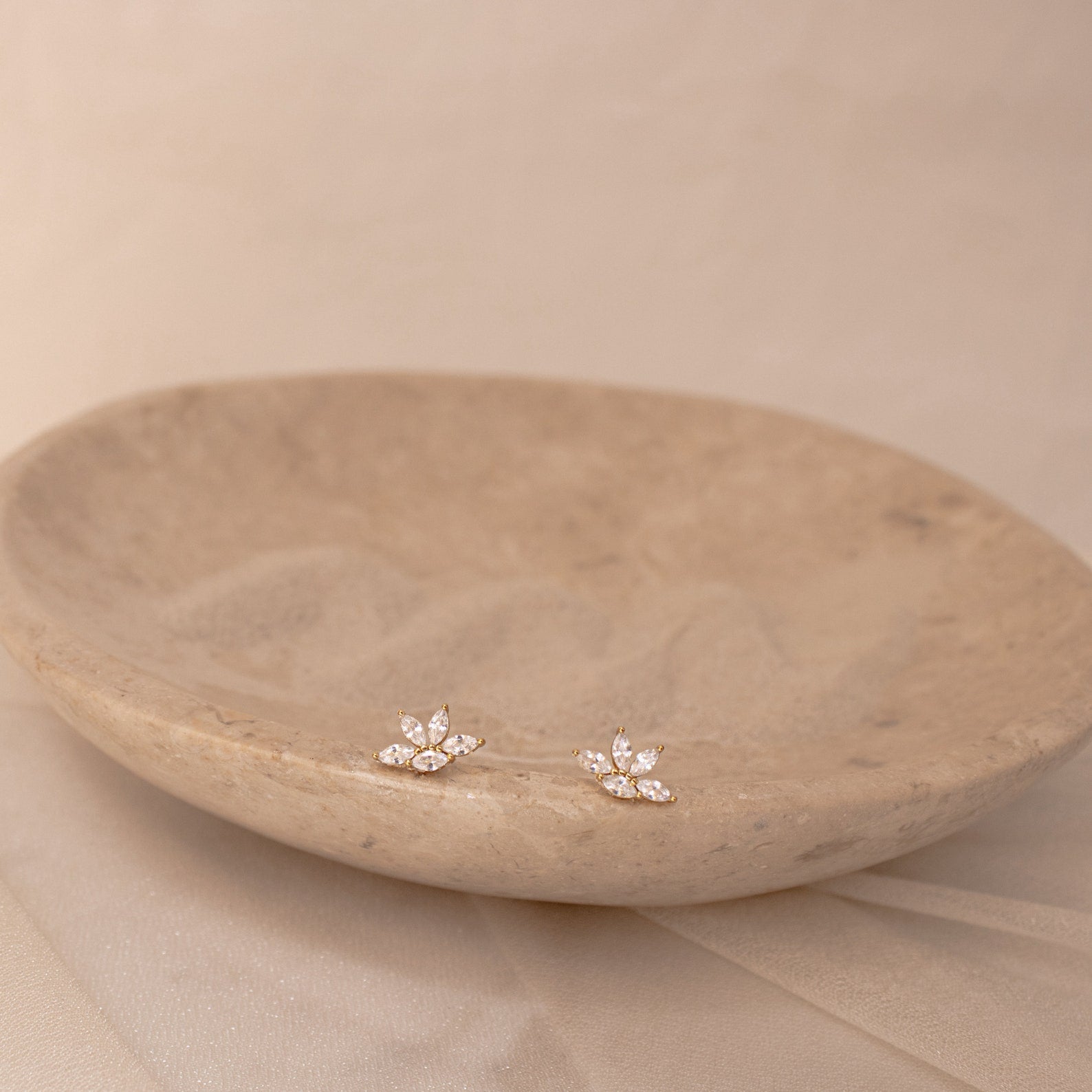 Marquise diamond stud earrings set in 18K gold, arranged in a delicate floral cluster on a textured beige dish.