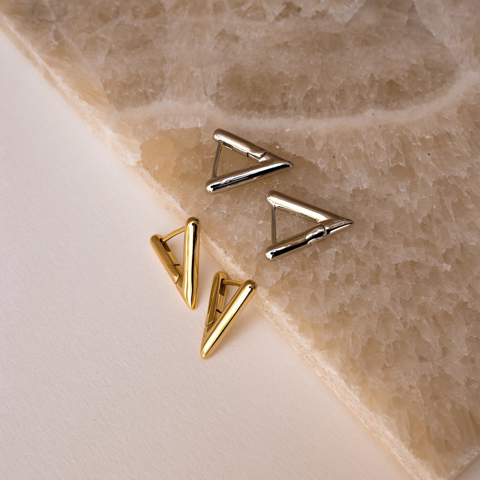 Two pairs of Triangle Huggies, in gold and silver finishes, are displayed on a textured stone surface—an ideal accessory for effortless summer style.