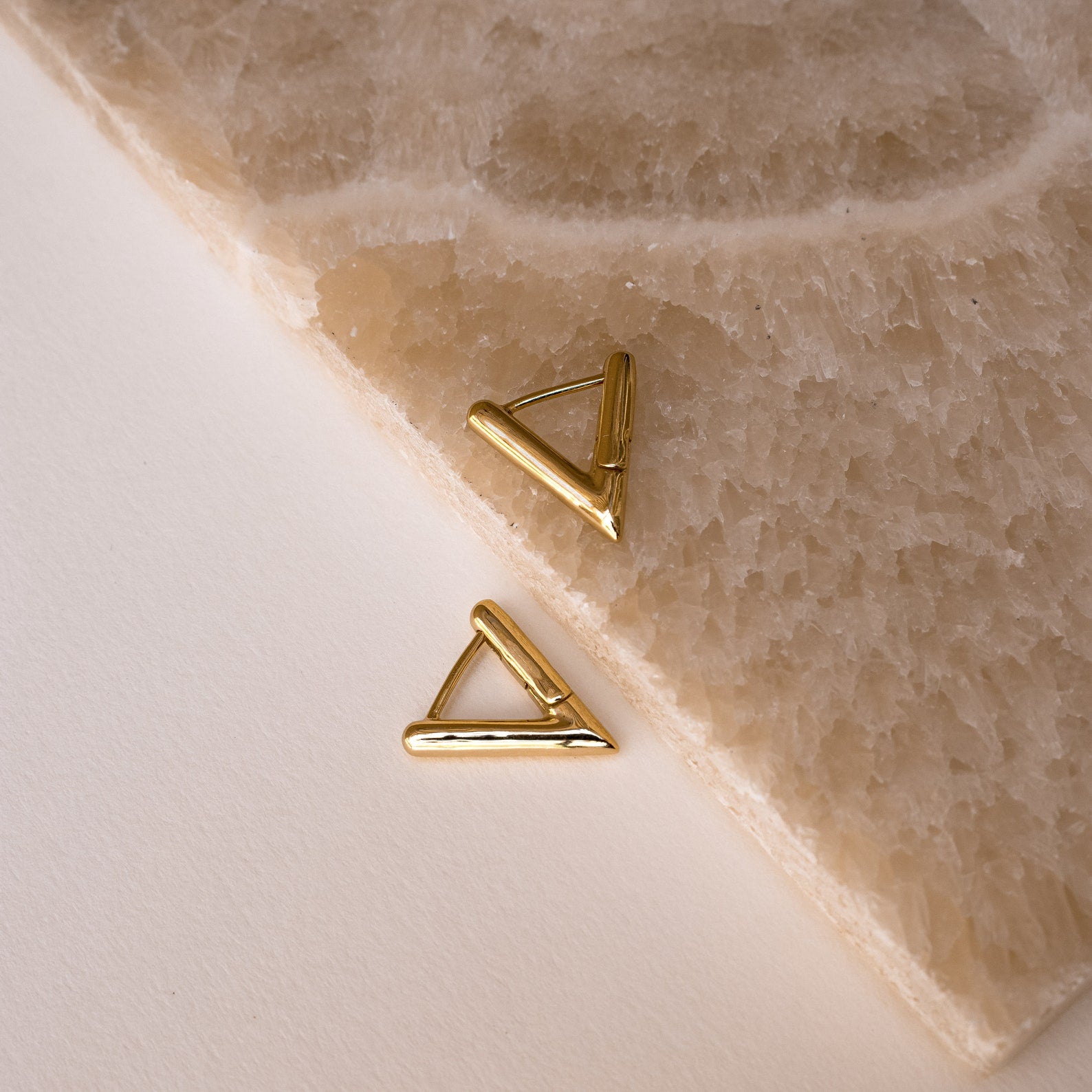 Two Triangle Huggies rest on a textured beige stone and off-white background—geometric earrings designed to elevate your summer style.