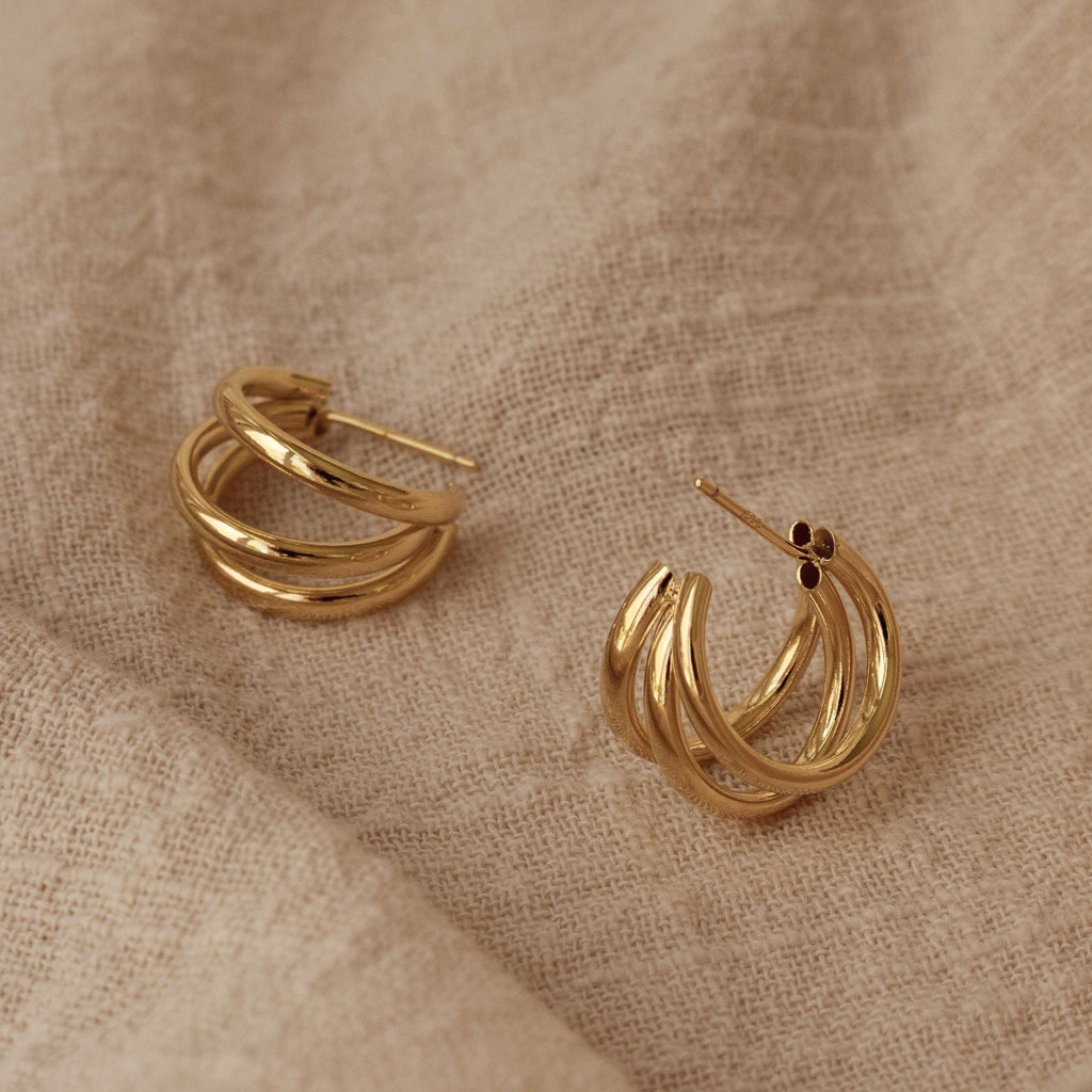 The Jenna Triple Hoops, minimalist gold earrings, rest elegantly on a beige textured fabric background, highlighting their sleek and modern design.