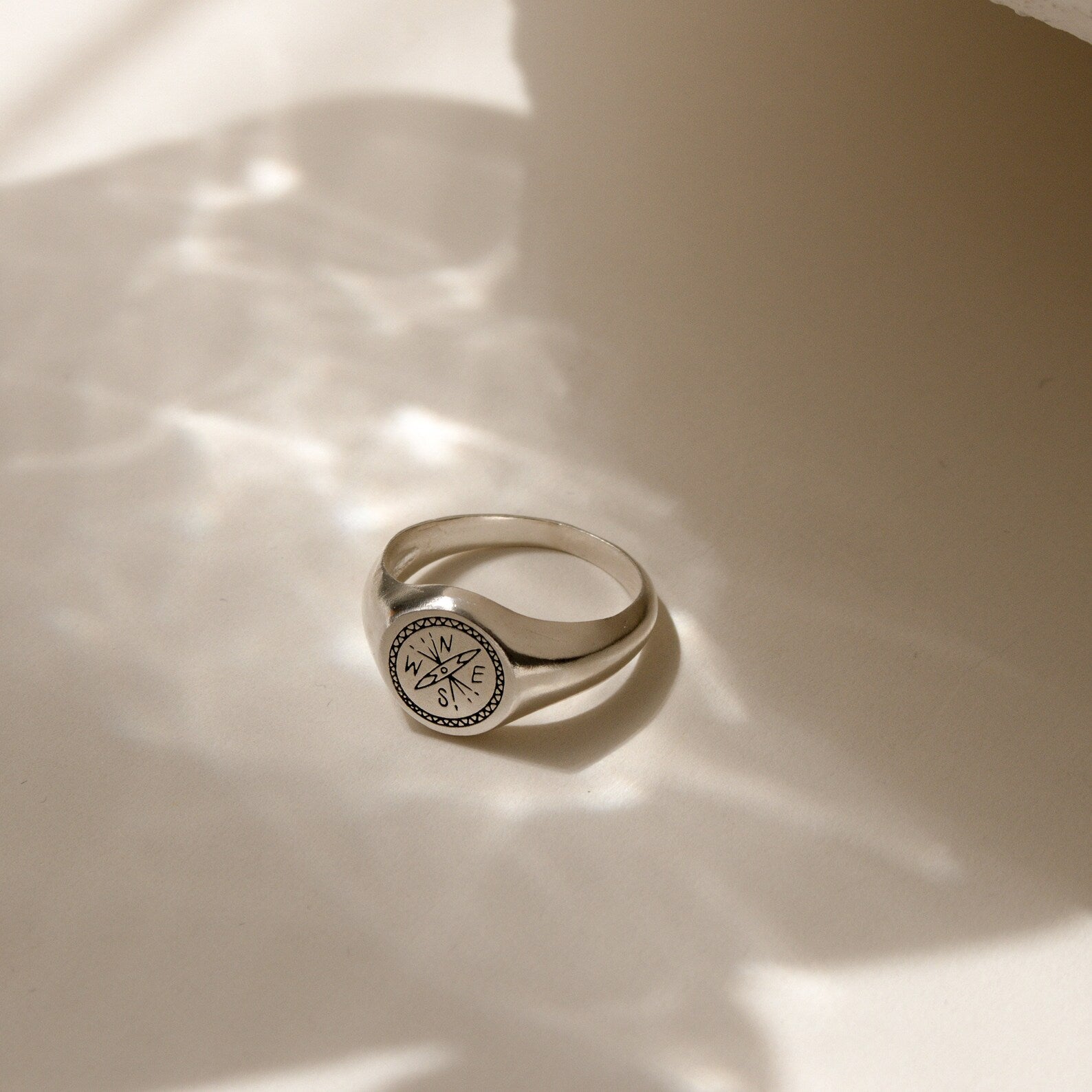 The Wanderlust Signet Ring features a silver compass design and rests on a light surface with soft shadows, embodying the spirit of adventure in elegant jewelry.