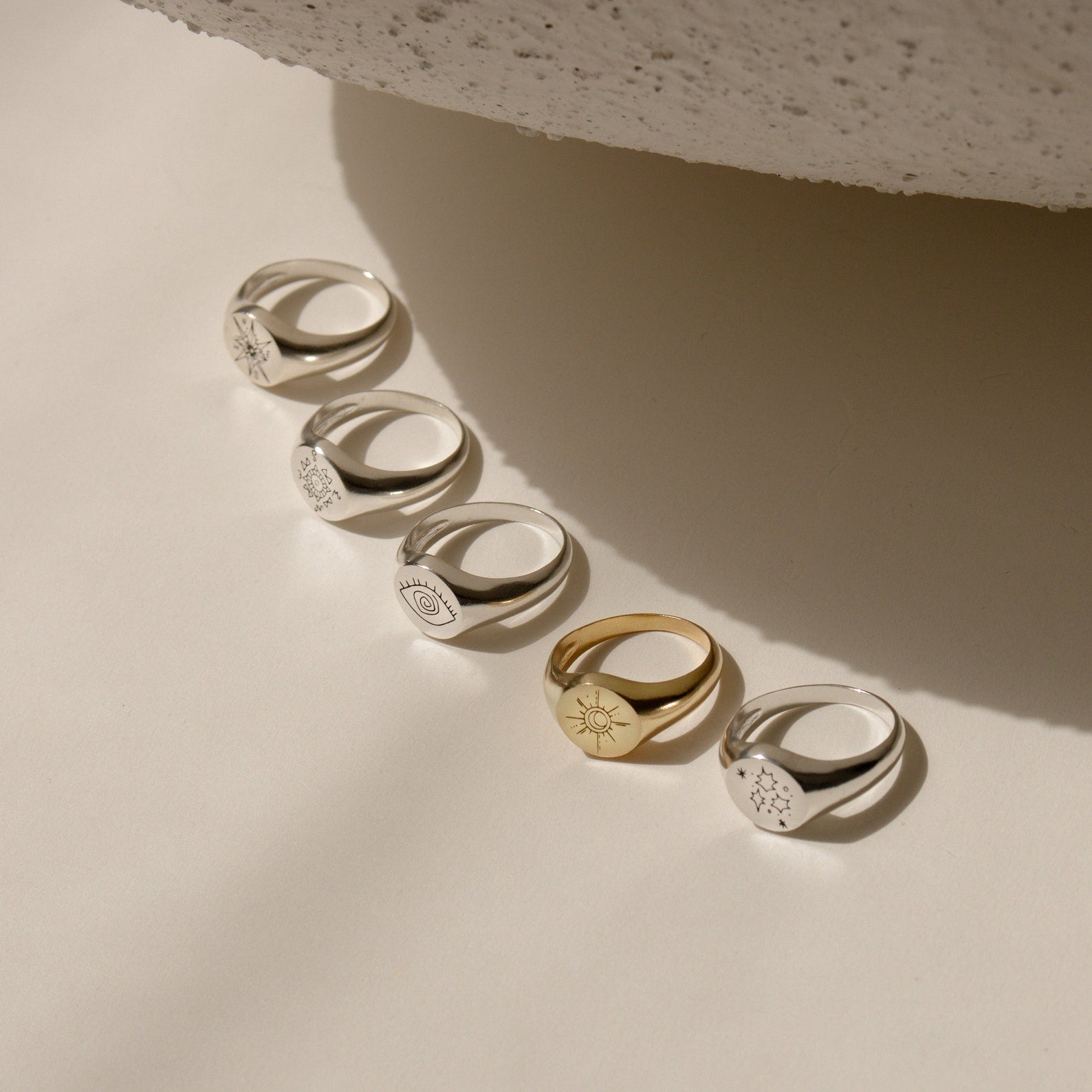 The Wanderlust Signet Ring, along with four other engraved rings—four in silver and one in gold—are elegantly displayed on a neutral surface under soft light, highlighting their timeless appeal.