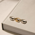 The Wanderlust Signet Rings—two in silver, one in gold—are displayed atop a large closed beige book under soft lighting, highlighting their elegant design.