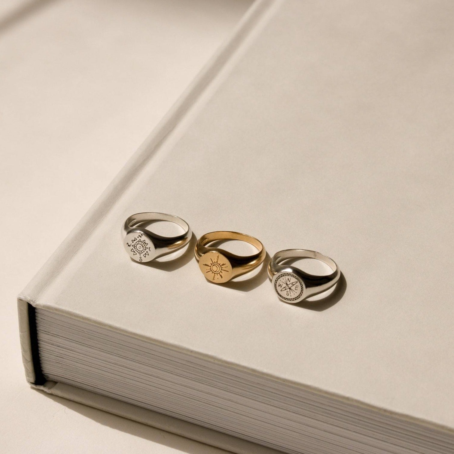 The Wanderlust Signet Rings—two in silver, one in gold—are displayed atop a large closed beige book under soft lighting, highlighting their elegant design.