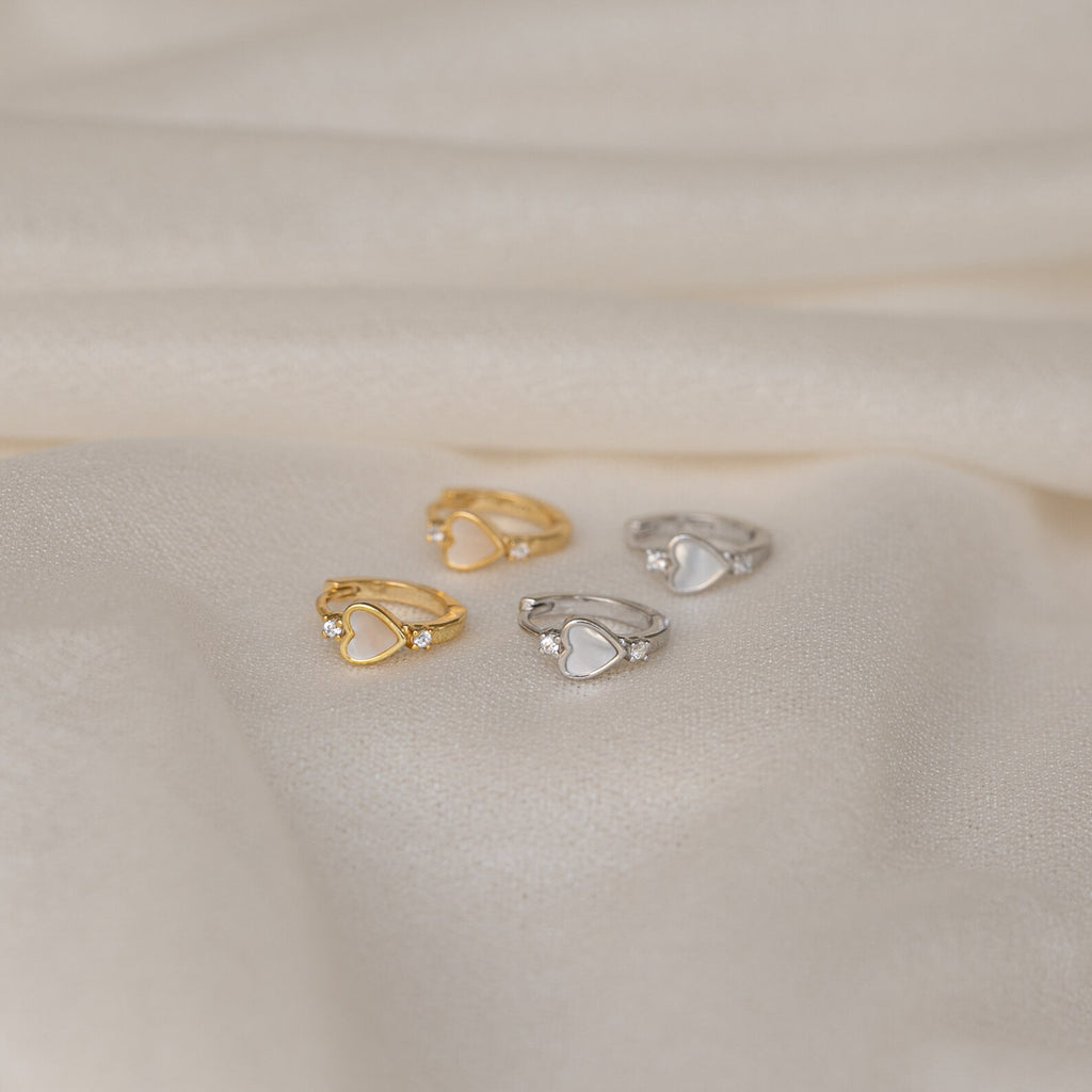 Four heart-shaped rings, two gold and two silver, rest on soft cream fabric, echoing the timeless charm of Scarlett Pearl Huggies and capturing the elegance found in classic mother of pearl heart jewelry.