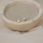 Scarlett Pearl Huggies are shown shimmering beside gold heart-shaped stud earrings on the rim of a round marble bowl filled with sand.