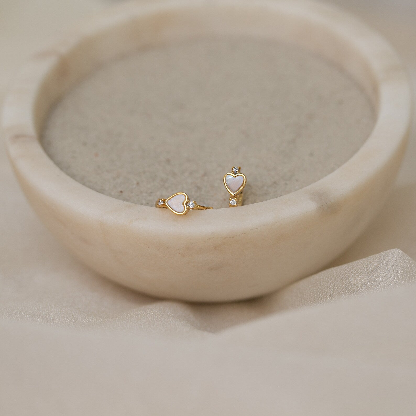 Scarlett Pearl Huggies are shown shimmering beside gold heart-shaped stud earrings on the rim of a round marble bowl filled with sand.