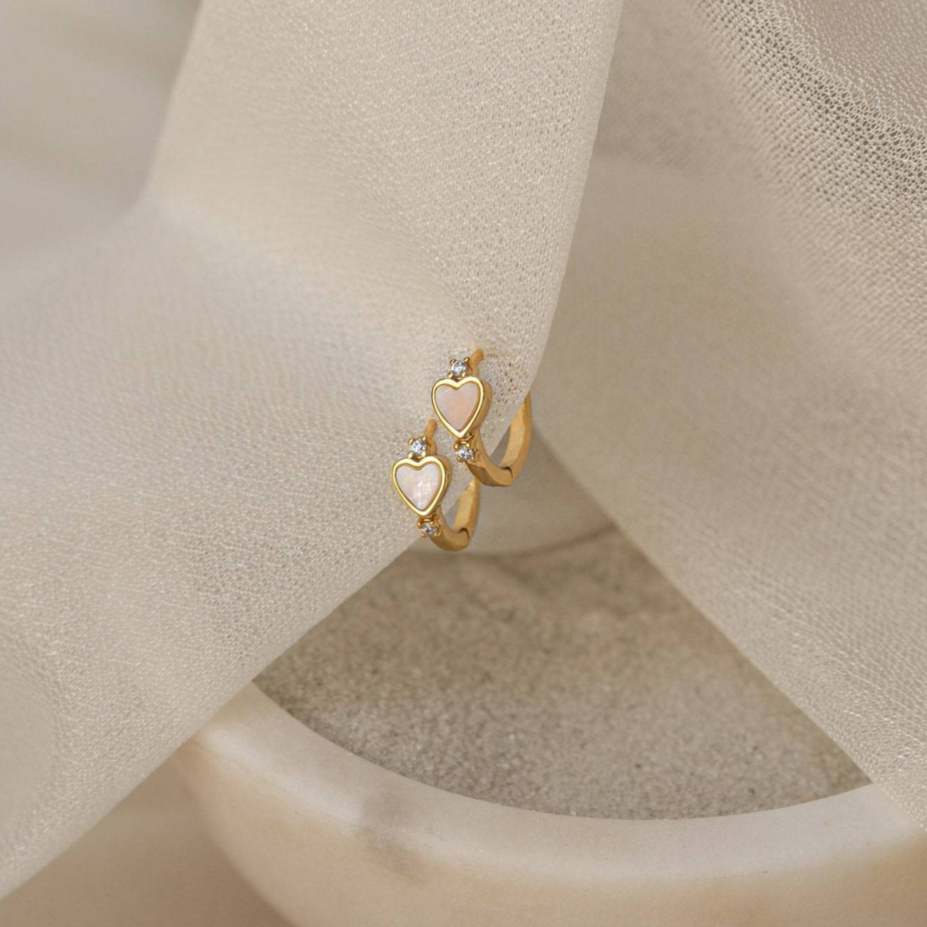 Scarlett Pearl Huggies, gold rings with pink heart-shaped charms and gems, and diamond huggie earrings are displayed on soft beige fabric and a stone dish.