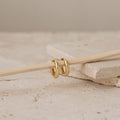 The Madison Hoops—two small classic hoops—hang elegantly on a thin wooden stick, set atop beige stone tiles. This minimalist piece is perfect for anyone who loves subtle, timeless jewelry.