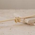 The Madison Hoops—two small classic hoops—hang elegantly on a thin wooden stick, set atop beige stone tiles. This minimalist piece is perfect for anyone who loves subtle, timeless jewelry.