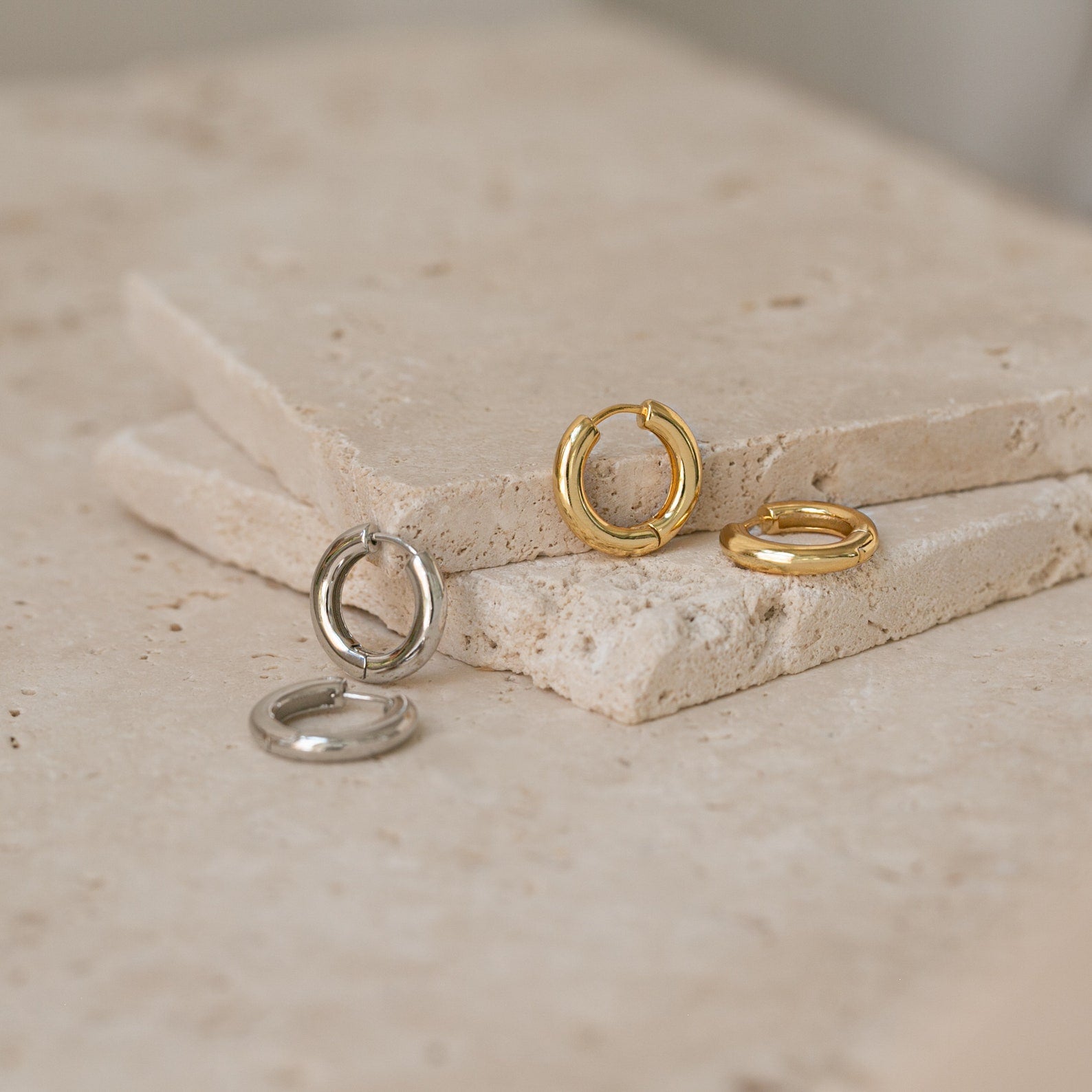 Two pairs of Madison Hoops, one gold and one silver, displayed on stacked stone slabs for a minimalist touch.