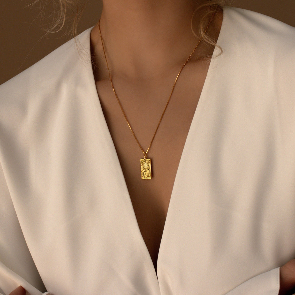 Woman in a white v-neck top wears the Tarot Necklace in Box Chain, a gold pendant necklace featuring an engraved rectangular charm inspired by the Major Arcana.