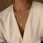 Woman in a white v-neck top wears the Tarot Necklace in Box Chain, a gold pendant necklace featuring an engraved rectangular charm inspired by the Major Arcana.
