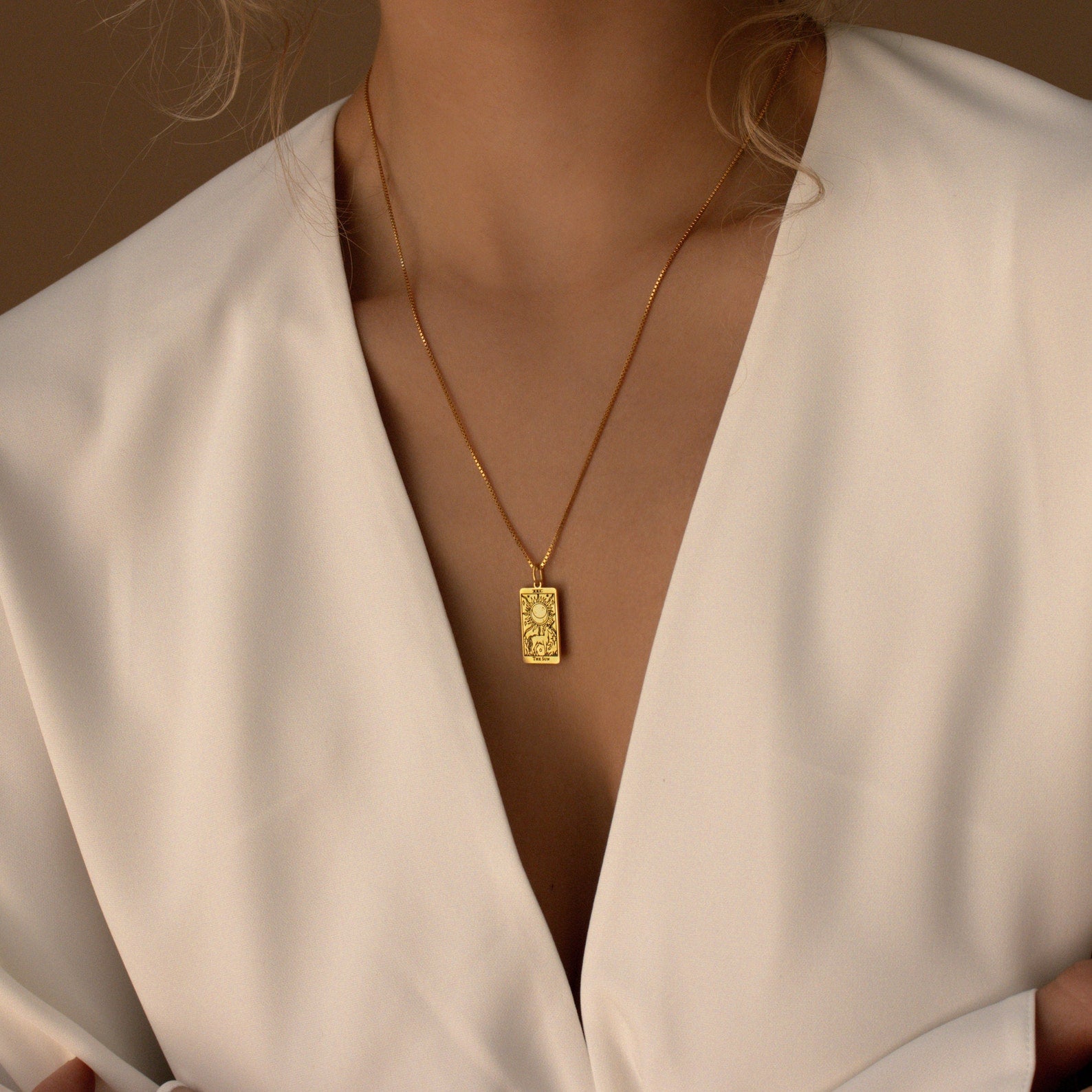 Woman in a white v-neck top wears the Tarot Necklace in Box Chain, a gold pendant necklace featuring an engraved rectangular charm inspired by the Major Arcana.