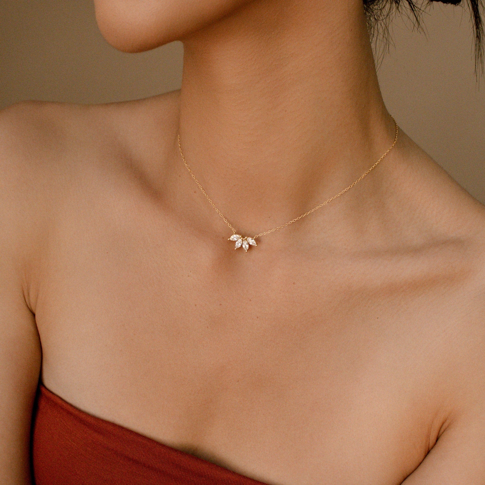 A minimalist shot of a woman’s neckline highlighting a delicate gold necklace with a trio of marquise diamonds in a symmetrical cluster design. The background is softly lit, and the model wears a strapless red top that complements the warm tones of the jewelry.