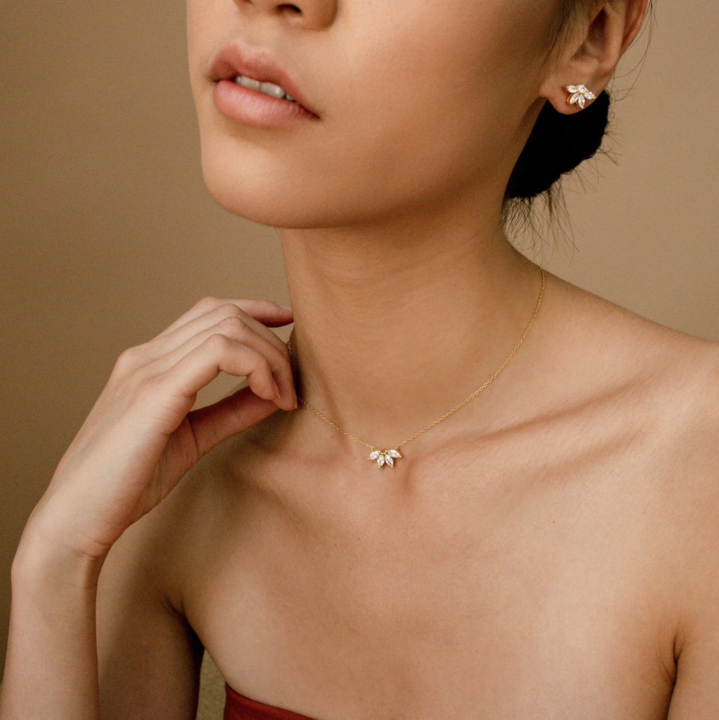 A stylish close-up of a woman wearing a matching jewelry set: a dainty gold necklace and stud earrings, both adorned with marquise-cut diamond clusters. Her hand gently touches her neck, emphasizing the elegant and feminine look of the coordinated set.
