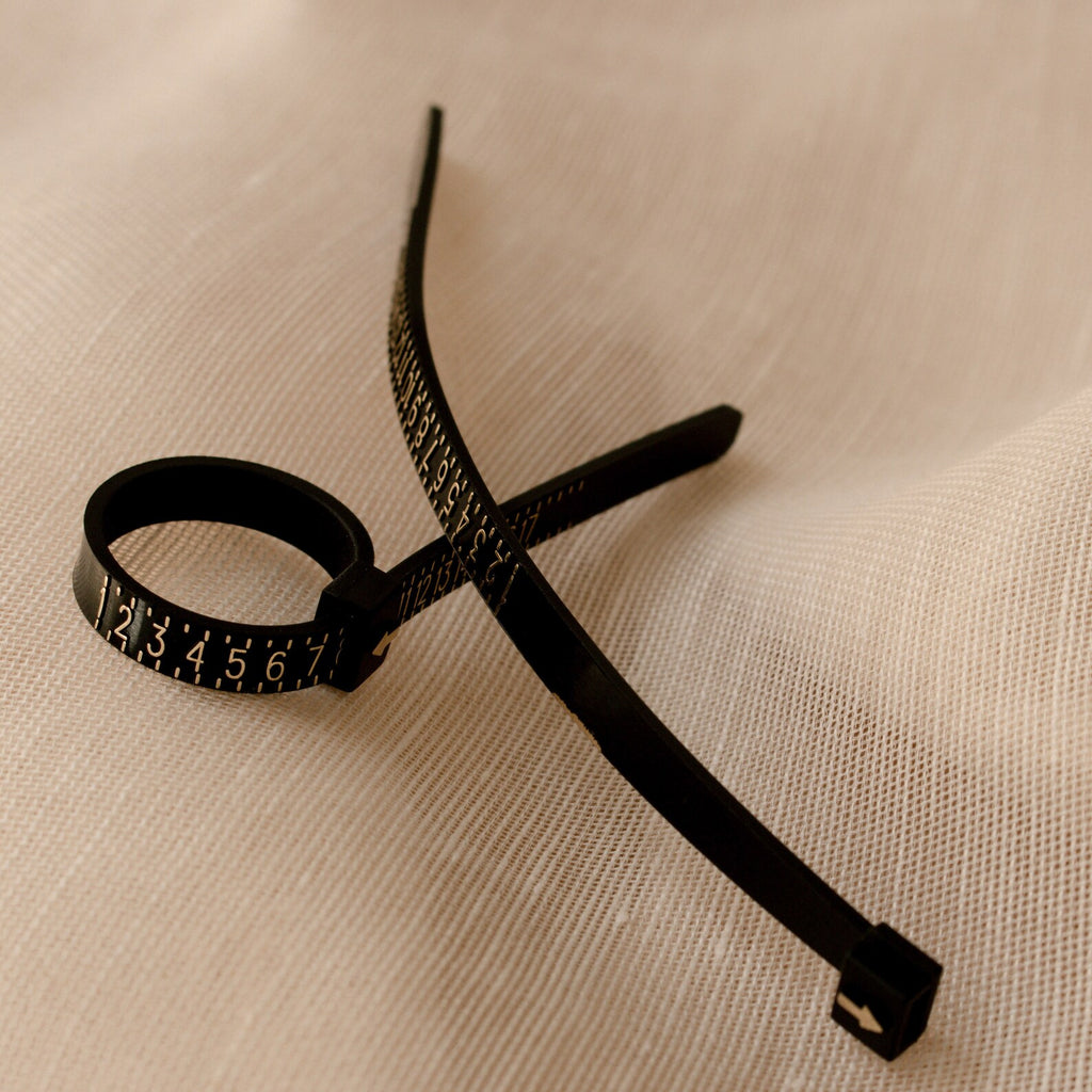 The Ring Sizer, a black cable tie with numbered markings for precise sizing, is placed on a textured beige fabric surface.