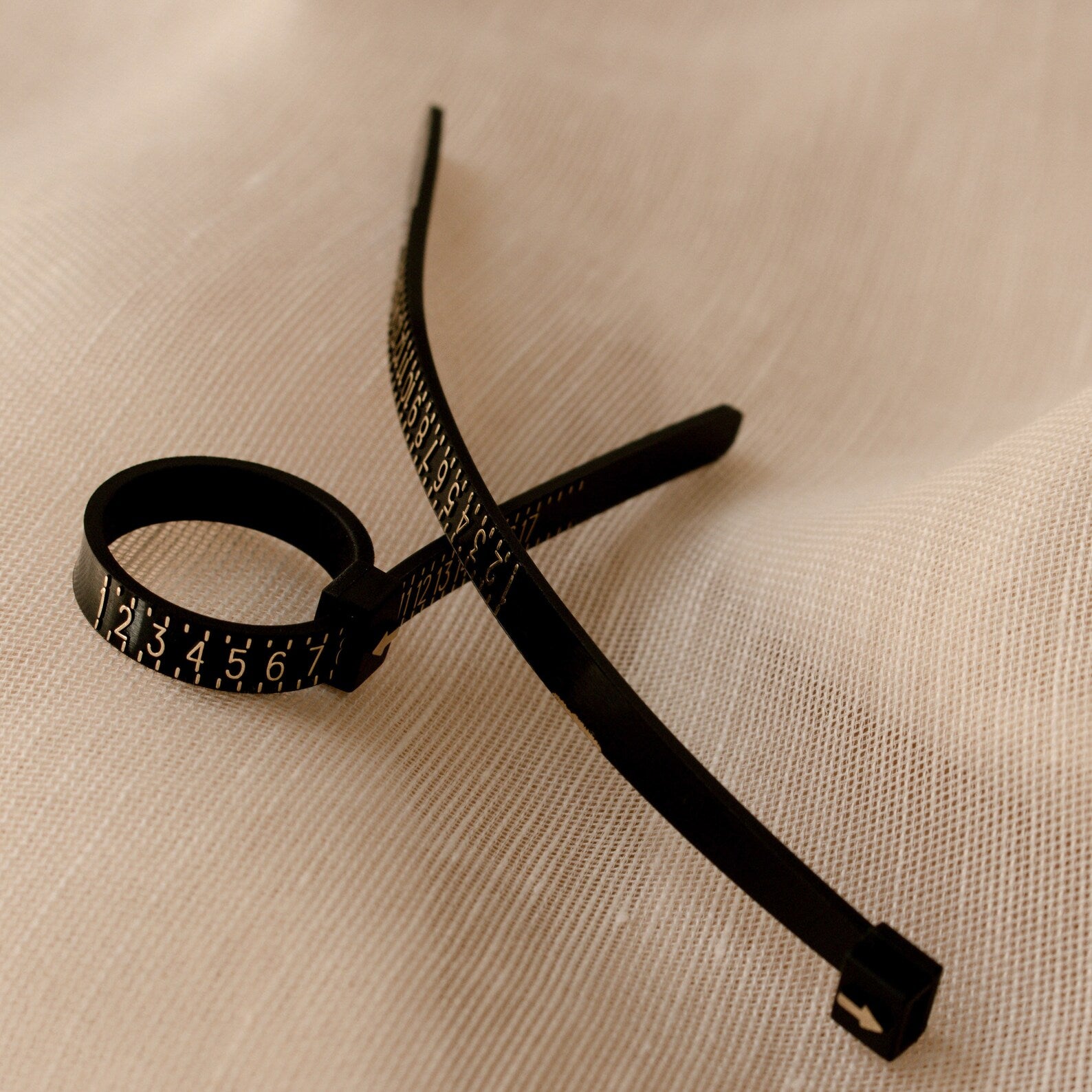 The Ring Sizer, a black cable tie with numbered markings for precise sizing, is placed on a textured beige fabric surface.
