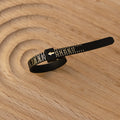 A black Ring Sizer with gold numbers and an arrow sits on a light wood surface with circular grooves, perfect for precise ring measurement.