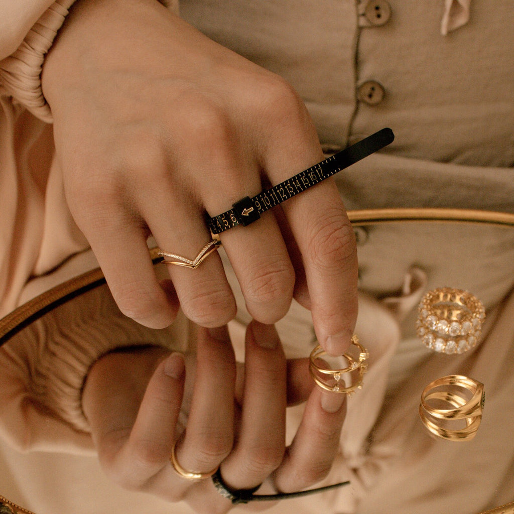 A hand adorned with gold rings uses the Ring Sizer for precise measurement, its reflection visible in a mirror scattered with additional rings.