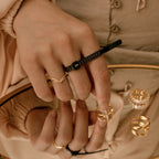 A hand adorned with gold rings uses the Ring Sizer for precise measurement, its reflection visible in a mirror scattered with additional rings.