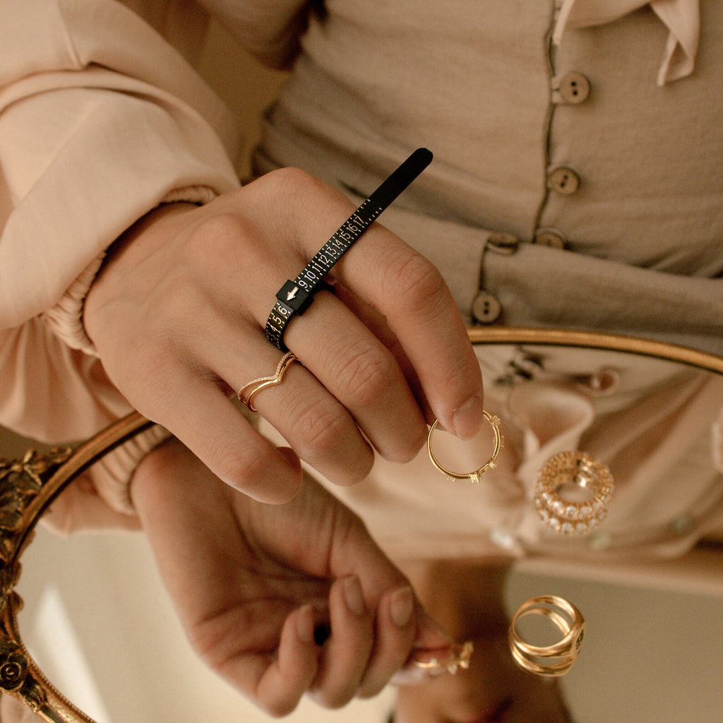 A hand uses the Ring Sizer to measure its ring size while wearing gold rings, reflected in a mirror.