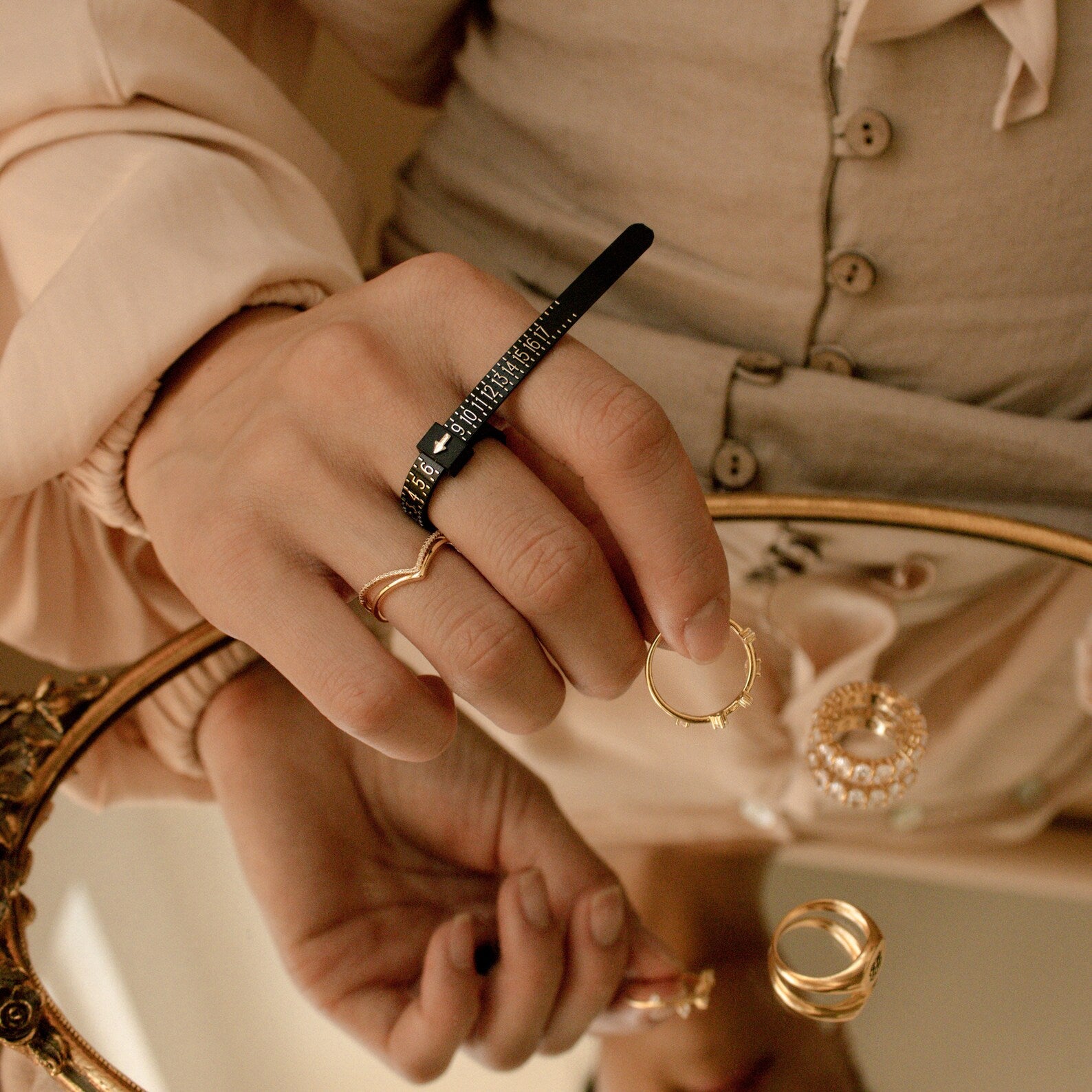 A hand uses the Ring Sizer to measure its ring size while wearing gold rings, reflected in a mirror.