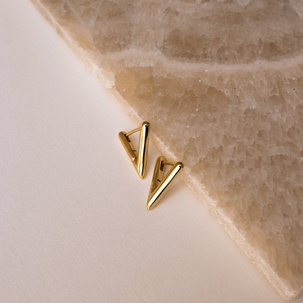 Triangle Huggies earrings with a gold geometric design rest on a beige surface beside a textured stone slab—an ideal accessory to elevate your summer look.