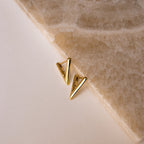 Triangle Huggies earrings with a gold geometric design rest on a beige surface beside a textured stone slab—an ideal accessory to elevate your summer look.