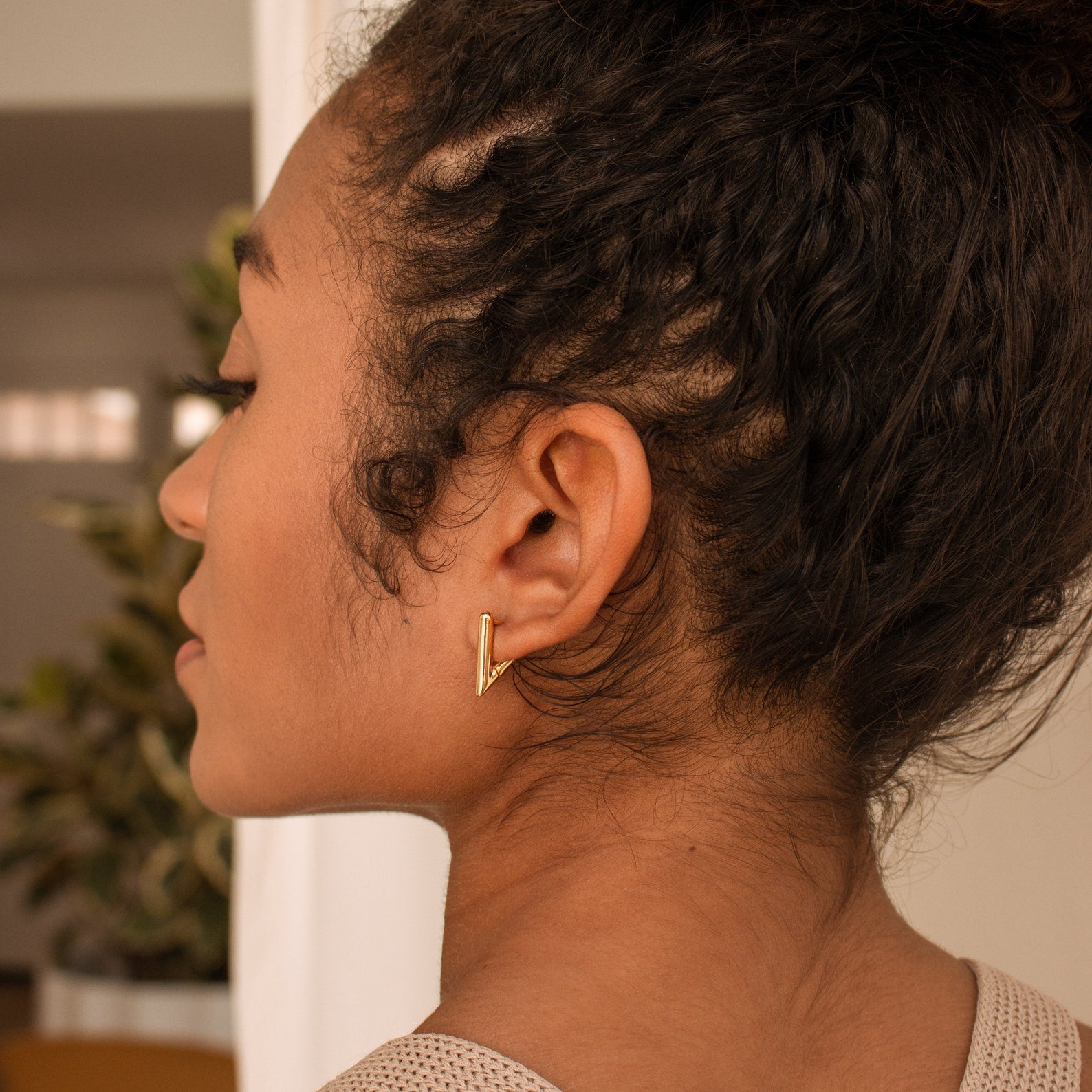 A woman with curly hair is shown in side profile wearing the Triangle Huggies, small gold geometric earrings, in her ear.