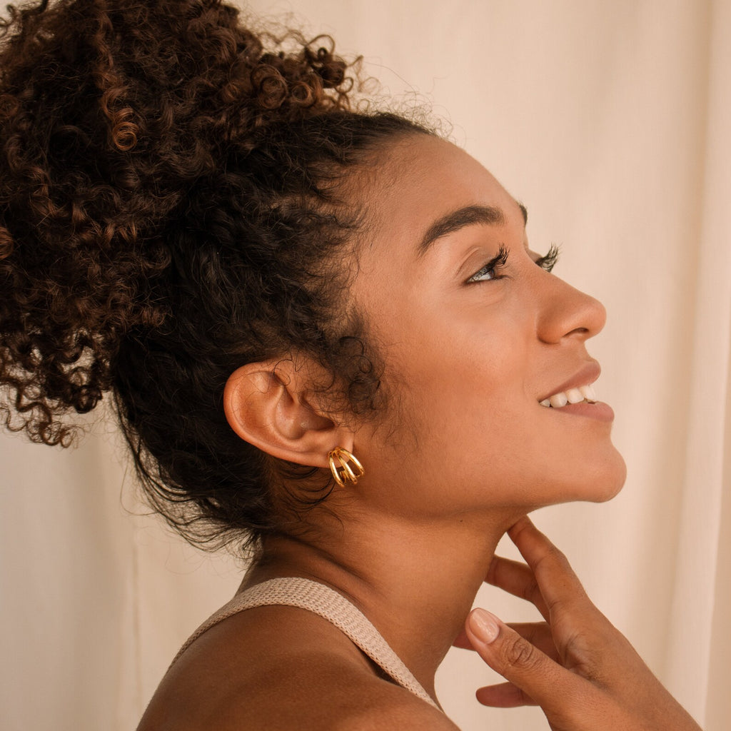 A woman with curly hair smiles, touching her neck, while wearing Jenna Triple Hoops against a soft, neutral background for a modern, minimalist look.