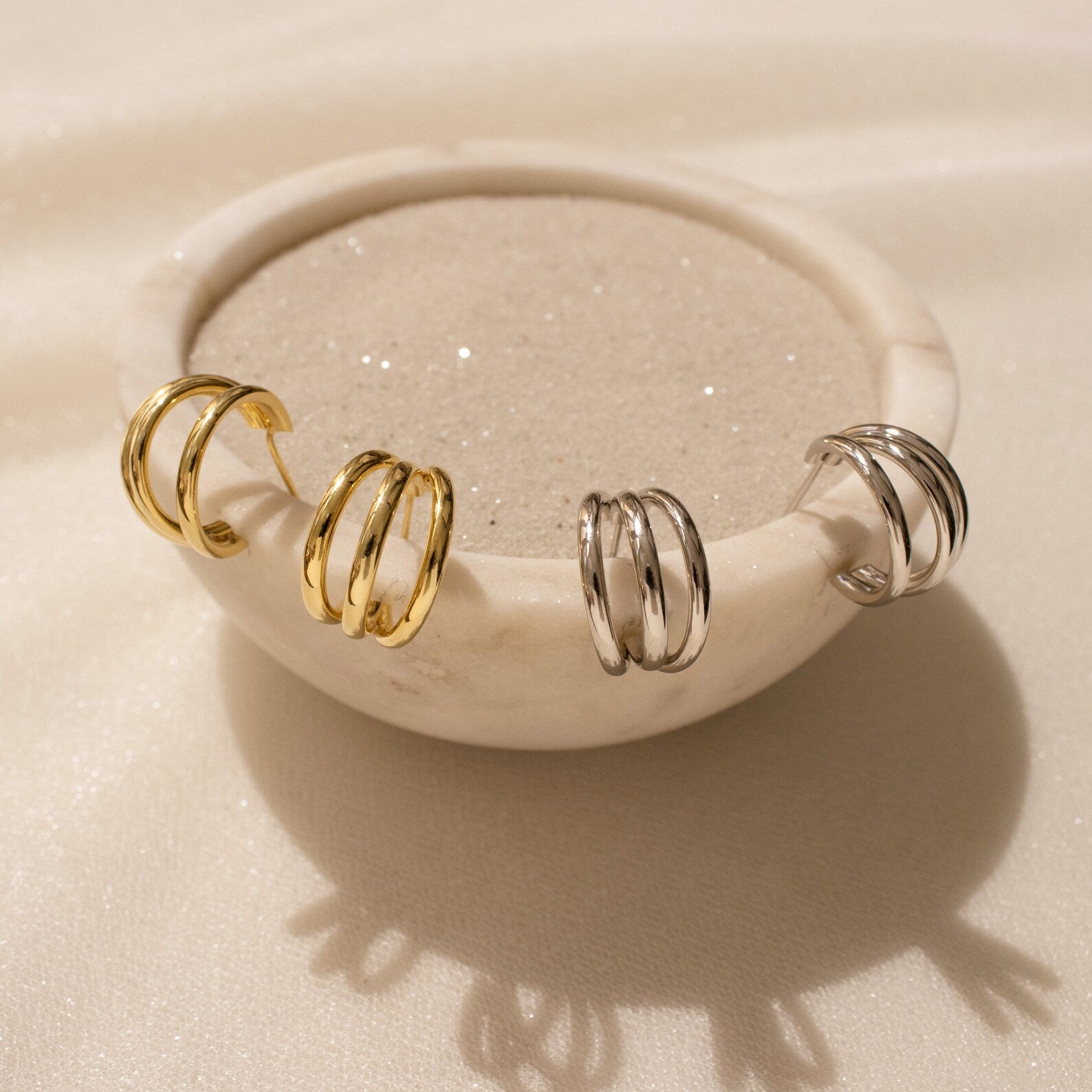 A marble bowl displays the Jenna Triple Hoops—modern earrings with gold and silver hoops—on its rim, embracing a minimalist aesthetic.